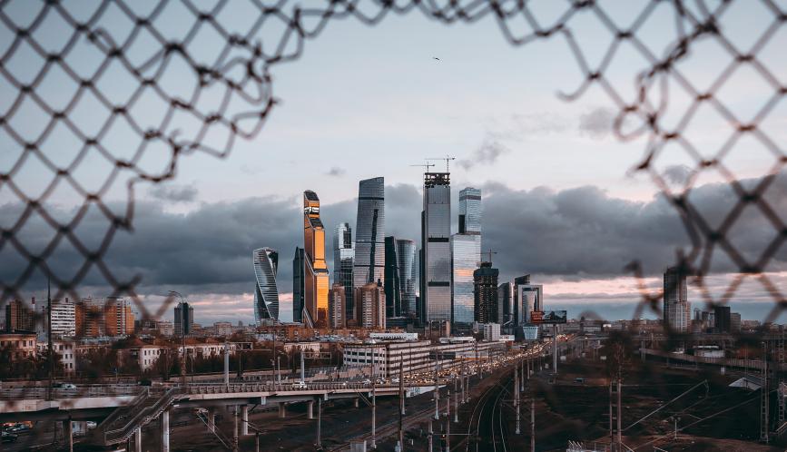 A view of the skyline of Moscow, as seen through a hole in a chain-link fence.