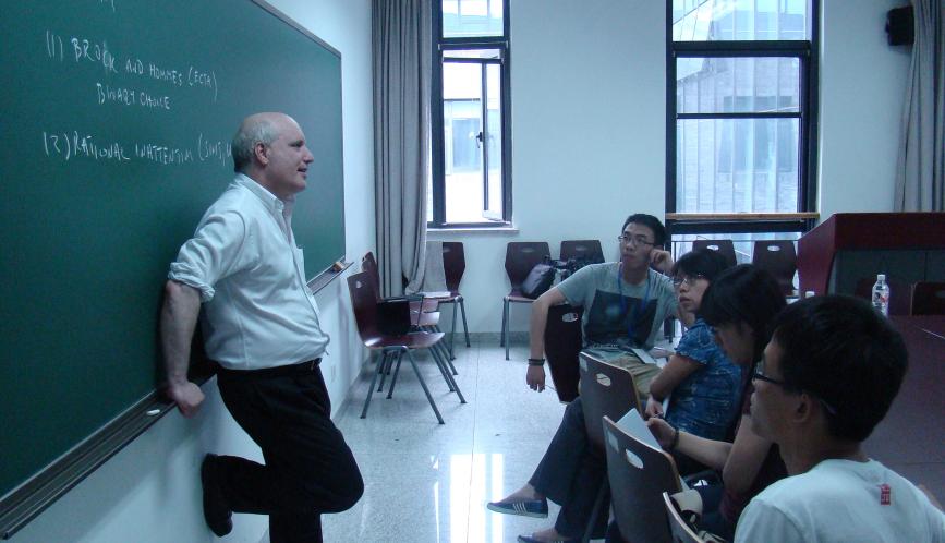 Steven Durlauf standing in front of a chalkboard, in conversation with a group of students sitting at desks in a classroom.