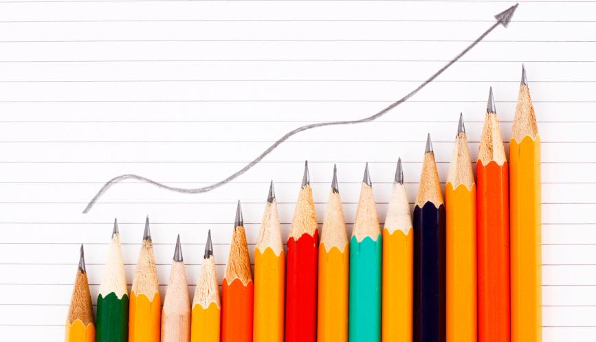 A stock image of different colored pencils, arranged in an escalating fashion, while a drawn arrow points upwards on a piece of paper behind them.