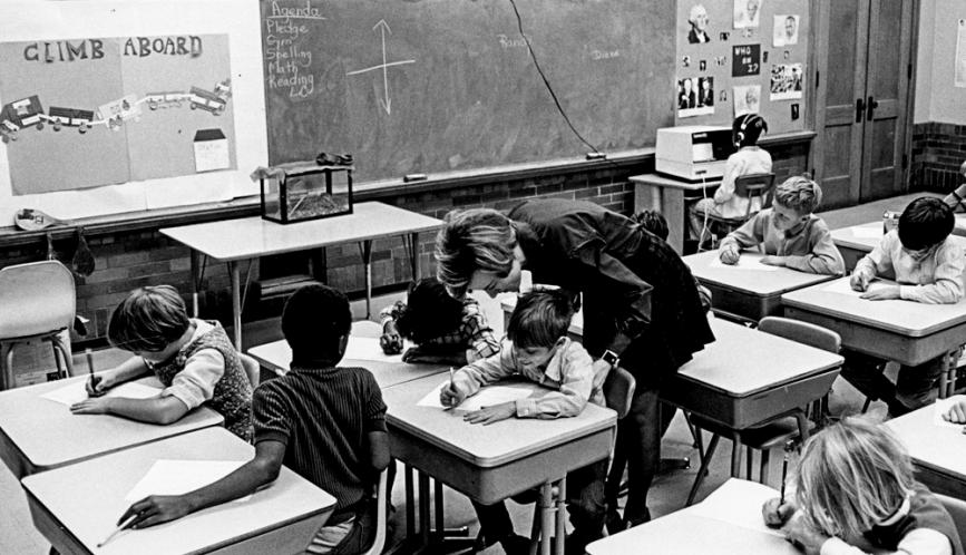 A black-and-white cover photo from the University of Chicago Photographic Archive, [apf4-01982], Special Collections Research Center, showing a female teacher bending over to assist a student at their desk, while other students in the classroom work independently.