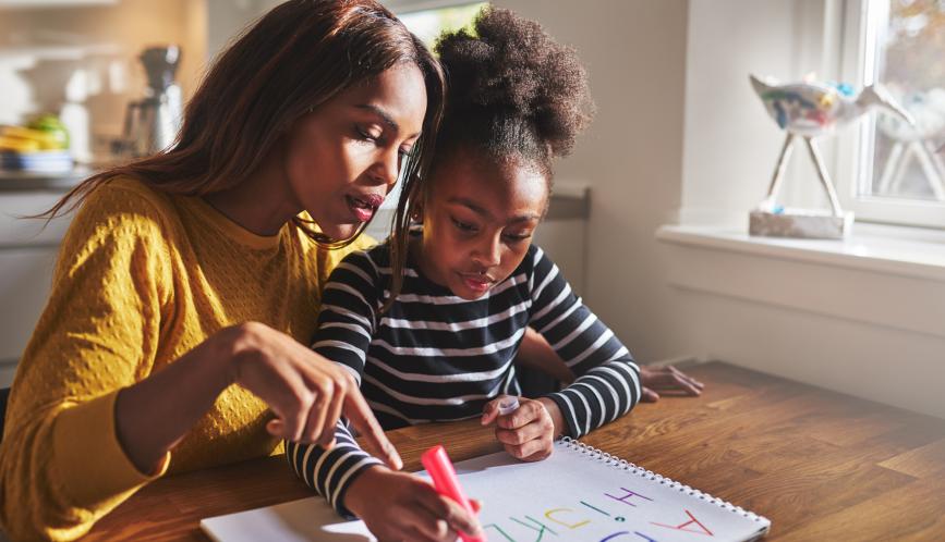 A stock image of a mother and daughter seated a table, the daughter is writing the alphabet in colorful marker.