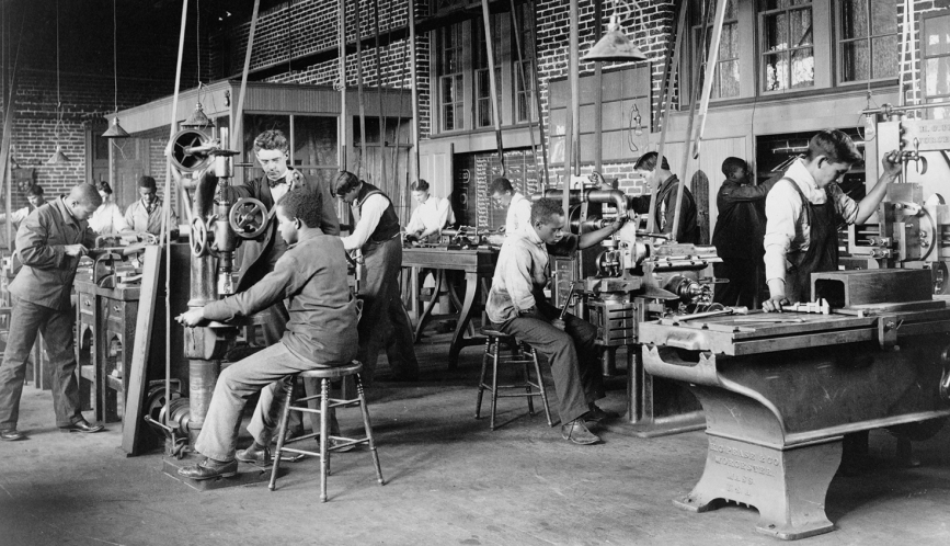 A historic, black and white image of a shop class at work at different machines.
