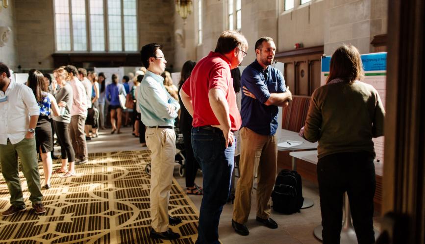 Many students in conversation during poster sessions, Jeff Smith is in the foreground looking at a student's poster.