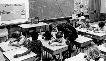 A black-and-white cover photo from the University of Chicago Photographic Archive, [apf4-01982], Special Collections Research Center, showing a female teacher bending over to assist a student at their desk, while other students in the classroom work independently.