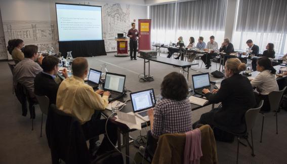 Tim Kautz presents at the front of the room, while a U-shaped table of attendees listens to him.